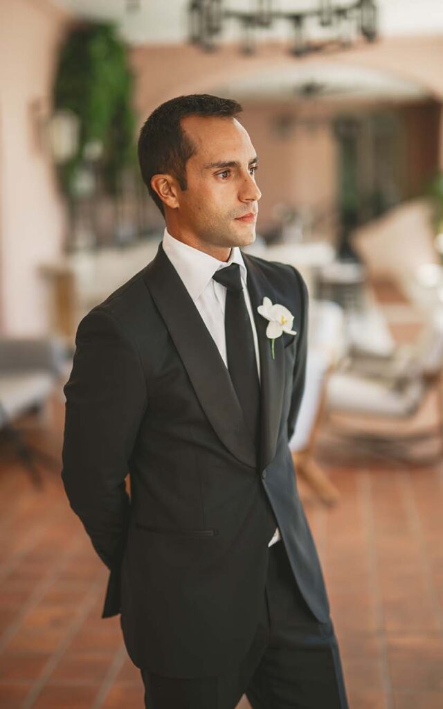 A groom in a black tuxedo standing in a courtyard.
