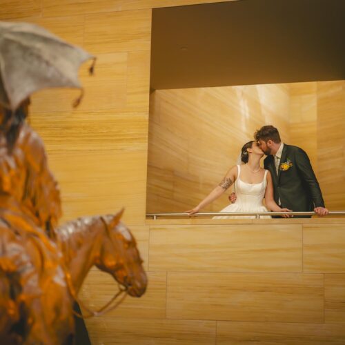 A bride and groom kiss on a balcony in a modern building, with a bronze statue of a rider on a horse visible in the foreground.