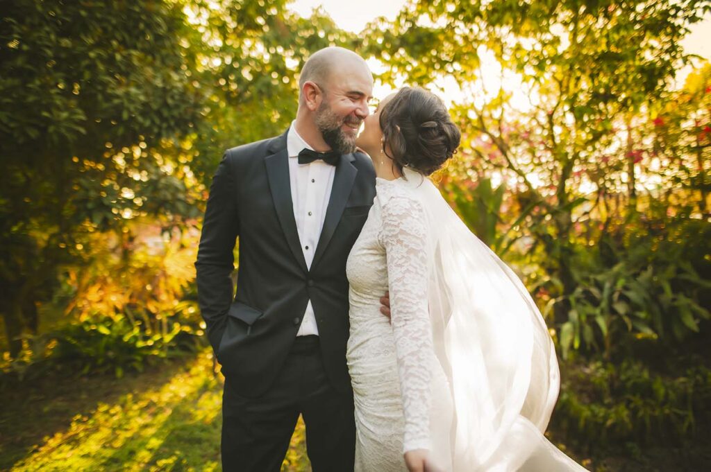 A bride in a white lace dress leans in to kiss a groom in a tuxedo as they stand outdoors in a sunlit garden.