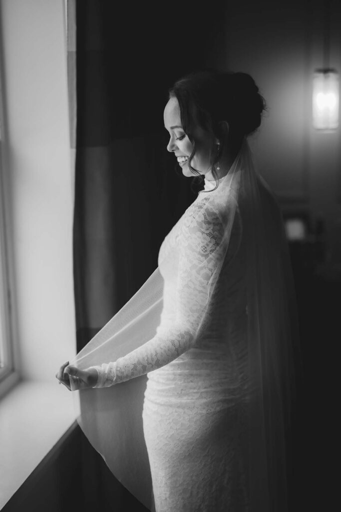 A woman in a lace wedding dress and veil stands by a window, looking down and smiling in a softly lit room.