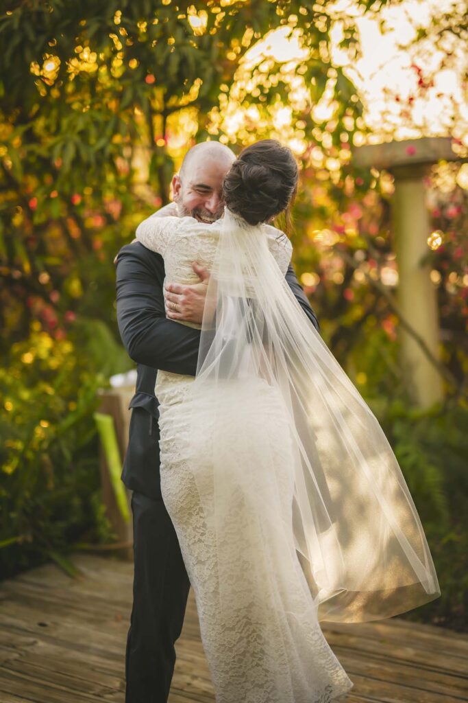 A bride and groom embrace outdoors on a wooden deck, with sunlight streaming through trees in the background.