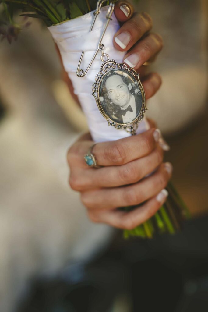 A person holds a bouquet wrapped in white ribbon, featuring a vintage photo of a man in a tuxedo framed in an ornate charm.