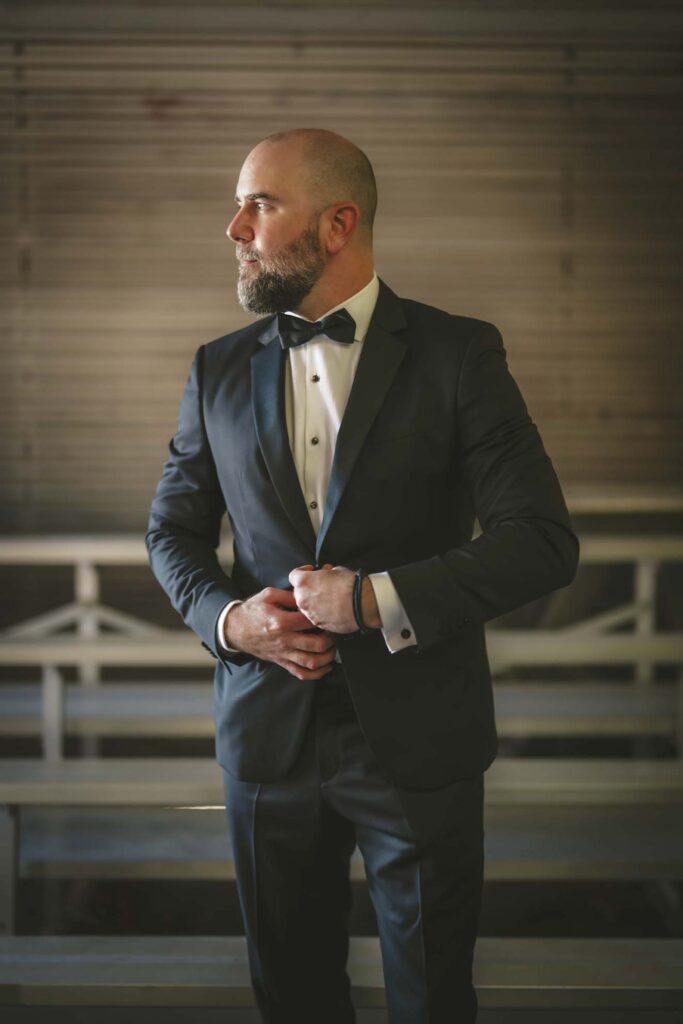 A man in a black tuxedo with a bow tie stands indoors, adjusting his cuff, and looks to his left.