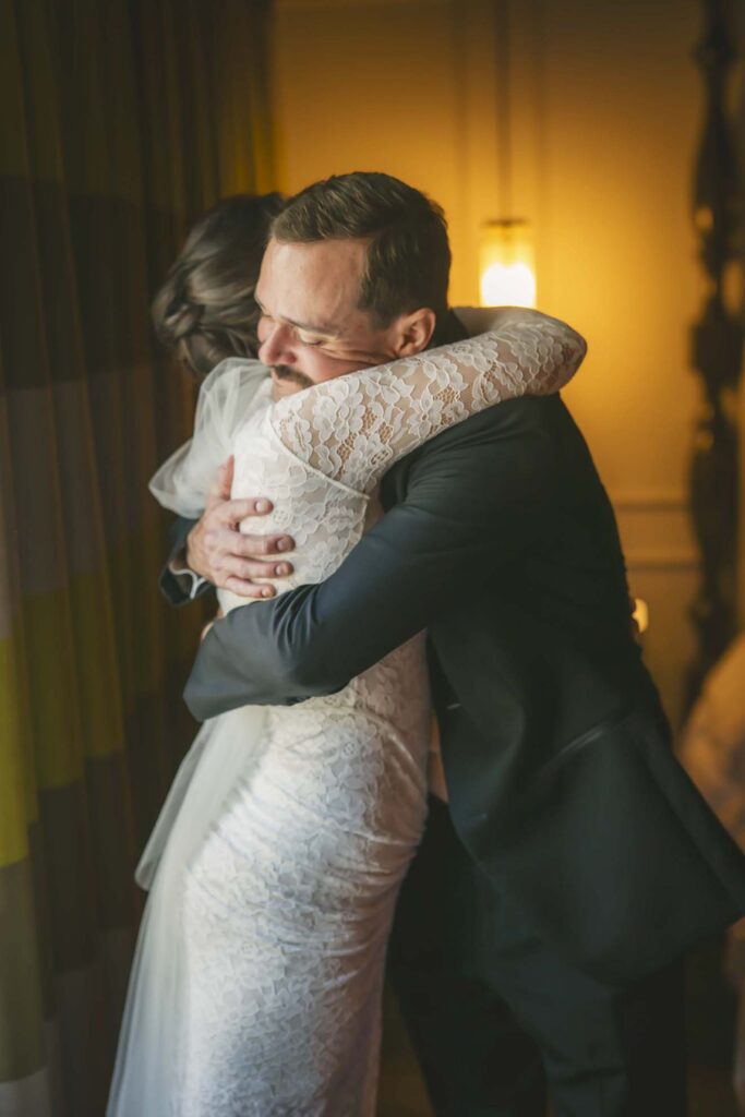A man in a suit and a woman in a lace wedding dress embrace closely in a warmly lit room.