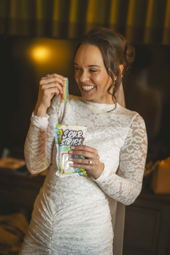 A woman in a white lace dress smiles while holding a bag of Sour Strips candy and lifting a piece out of the bag.