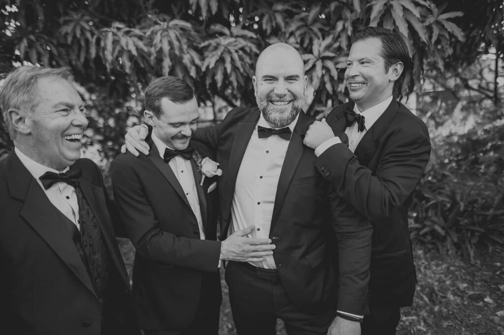 Four men in tuxedos stand together outdoors, smiling and laughing, with trees and foliage in the background.