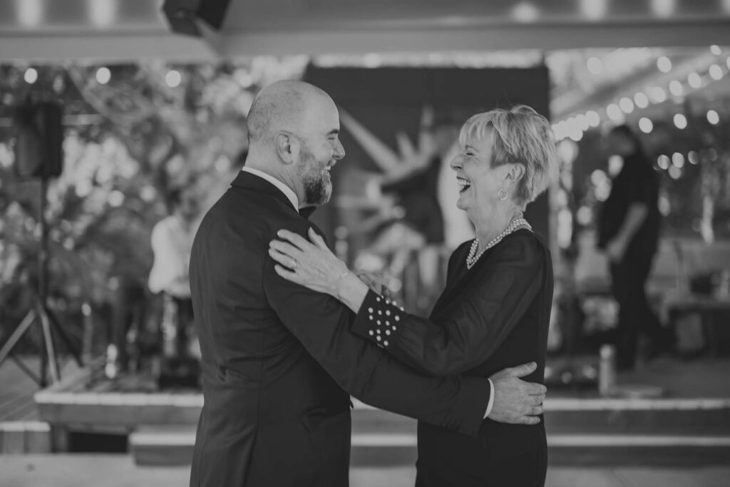 A man in a suit and a woman in a dress are smiling and dancing together in an indoor venue with string lights in the background.