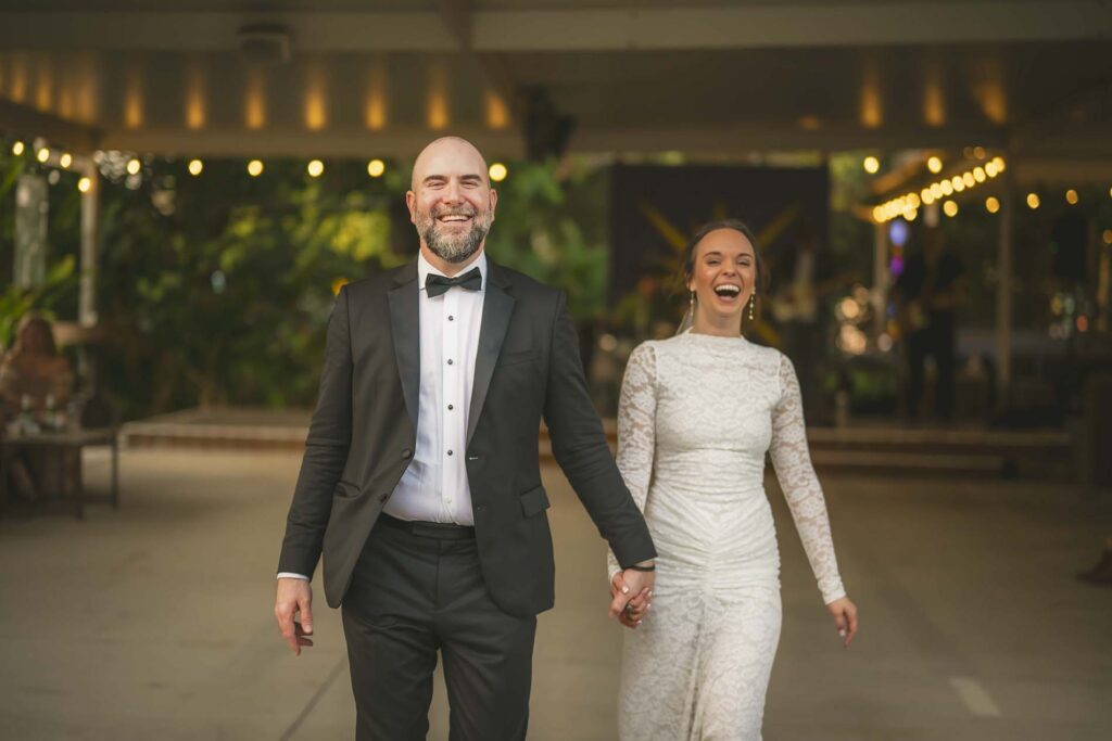A man in a tuxedo and a woman in a white dress walk hand-in-hand, both smiling, in an outdoor setting with string lights in the background.