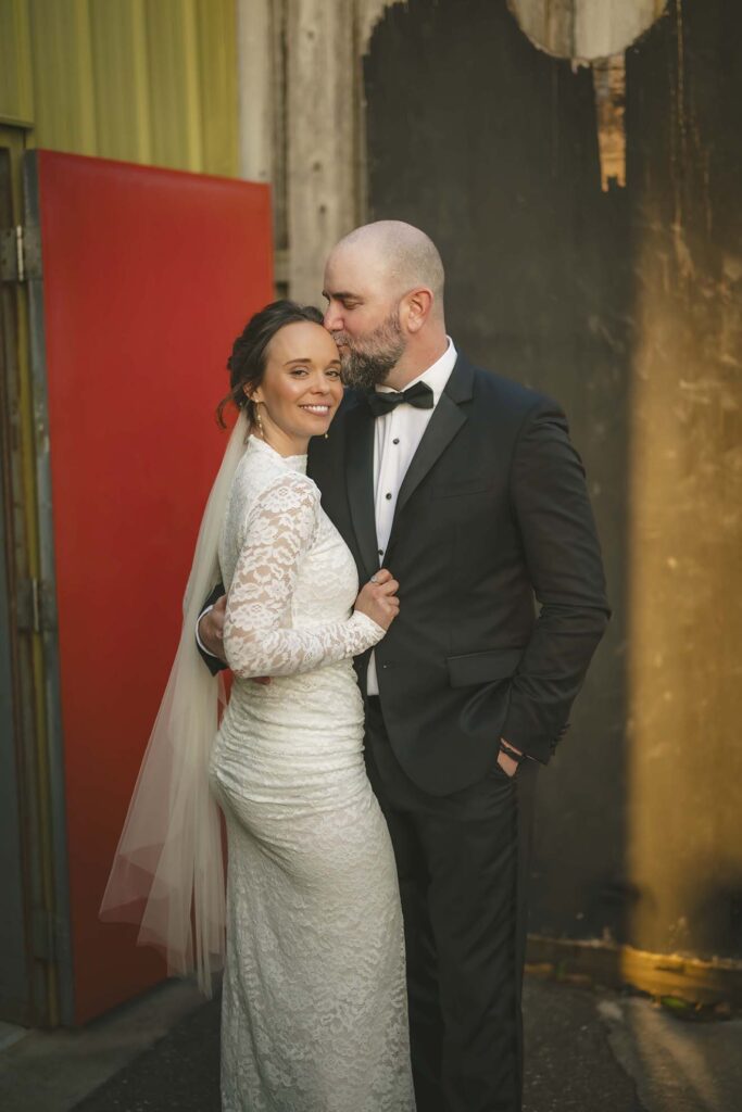 A bride in a lace dress and veil stands next to a groom in a black tuxedo; he kisses her forehead as they pose outdoors beside a red door and weathered wall.