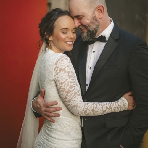 Bride and groom in formal attire embrace and smile with eyes closed, standing closely together in front of red and dark backgrounds.