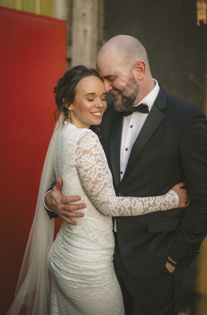 Bride and groom in formal attire embrace and smile with eyes closed, standing closely together in front of red and dark backgrounds.