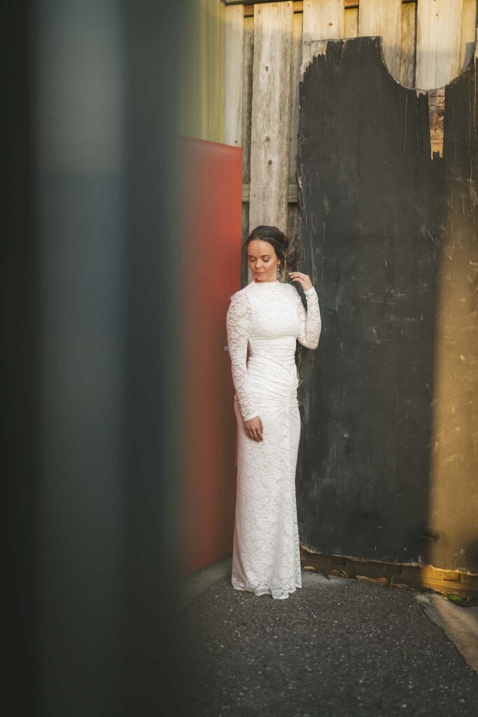 A woman in a long-sleeved white lace dress stands in an alleyway, looking down, with her hand touching her hair beside a weathered wooden fence and a red wall.