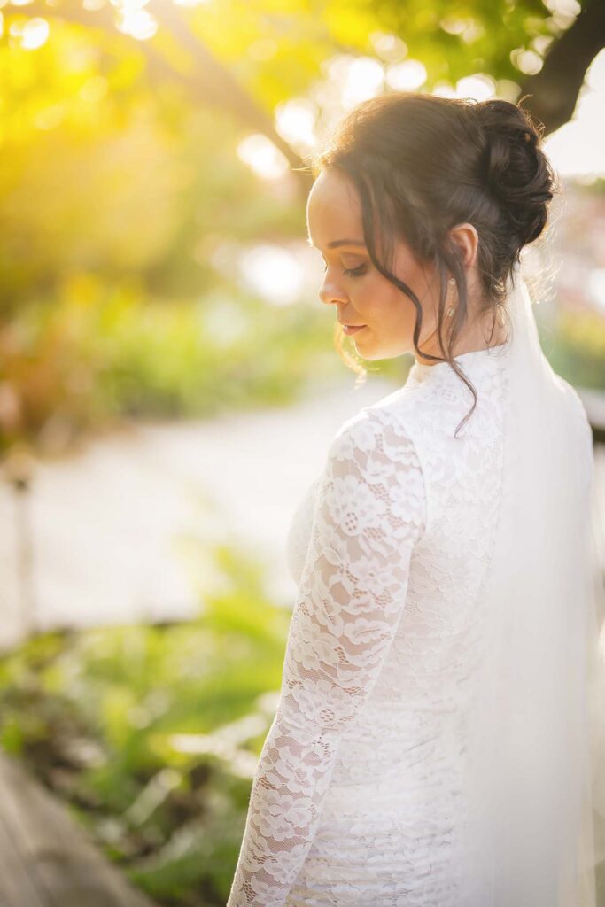 A woman in a white lace dress and veil stands outdoors, looking down, with sunlight filtering through trees in the background.