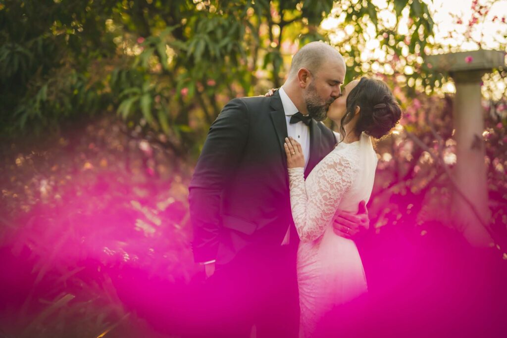 A couple in formal wedding attire shares a kiss outdoors, surrounded by greenery and bright pink foreground flowers.