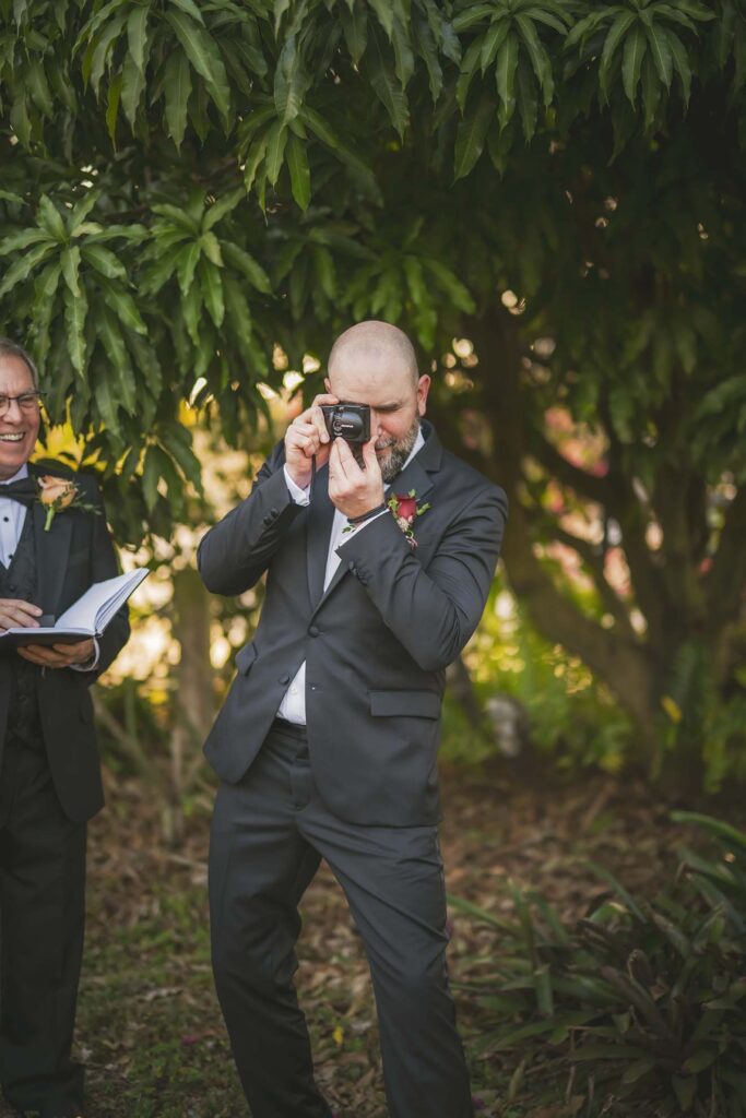 A man in a suit takes a photo with a camera outdoors, standing on grass near leafy trees, with another person in a tuxedo partially visible beside him.