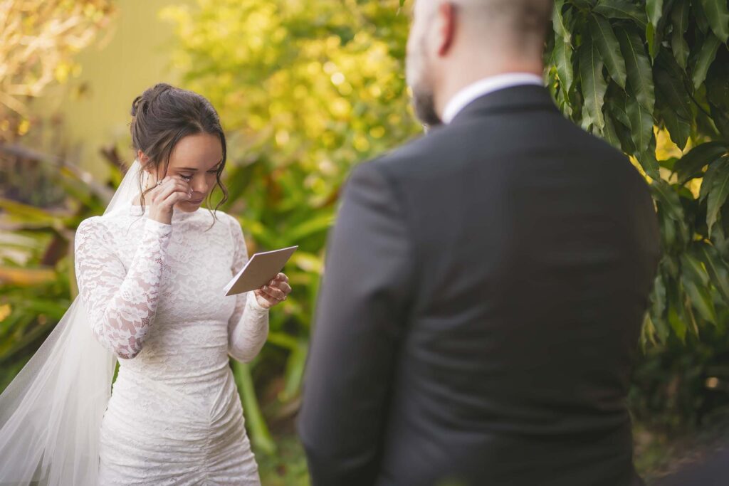 A bride in a white lace dress wipes away a tear while reading from a card, standing outdoors facing a man in a suit.