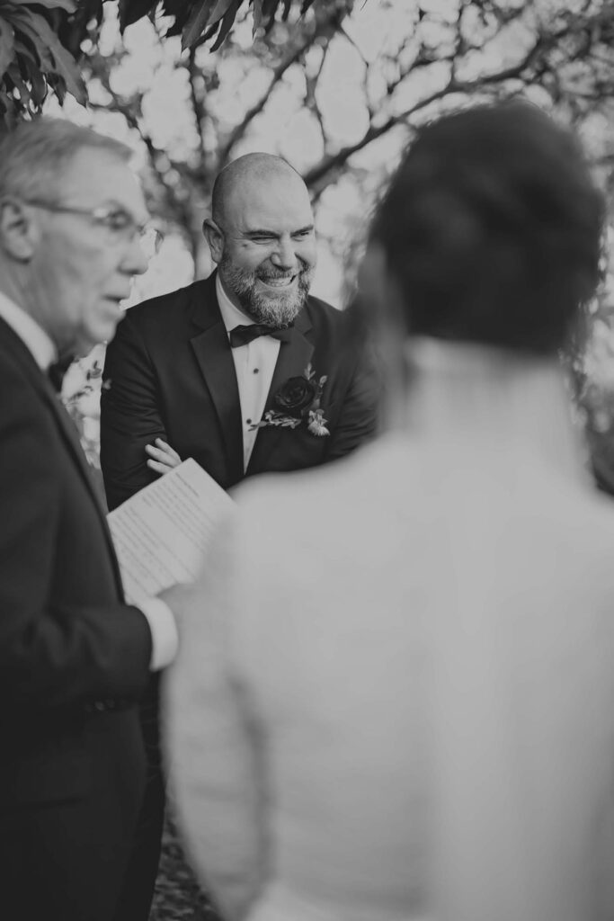 A man in a suit smiles during an outdoor ceremony, standing next to another man holding papers; a woman in a veil is in the foreground, seen from behind.