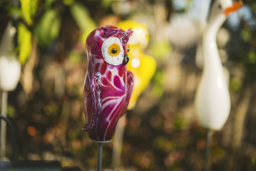 A pink and white glass owl ornament is displayed outdoors, with other bird-shaped ornaments blurred in the background.