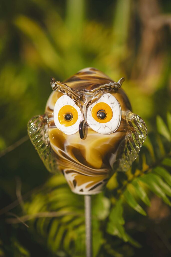 A glass owl figurine with yellow and brown swirls, orange eyes, and clear glass wings, mounted on a metal stick, set against a blurred green foliage background.