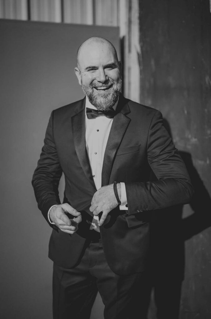 Man in a tuxedo smiles while buttoning his jacket, standing indoors against a plain and dark wall; the image is in black and white.