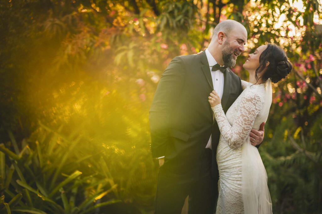 A bride in a lace dress and a groom in a black suit stand close together outdoors, smiling at each other in warm, golden sunlight.