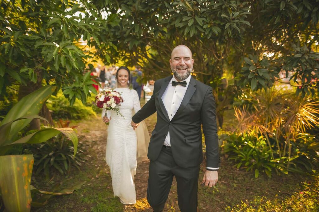 A man in a tuxedo and a woman in a white wedding dress hold hands and walk outdoors among green plants, with the woman carrying a bouquet.