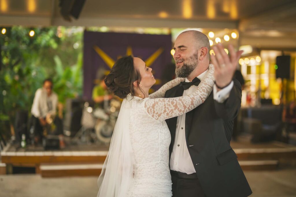 A bride and groom in formal attire dance together indoors, smiling and embracing, with a band performing in the background.