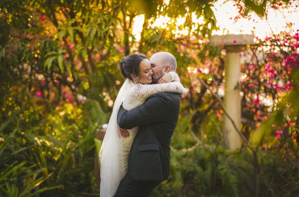 A groom in a dark suit lifts a bride in a white dress and veil as they embrace outdoors amid greenery and flowers at sunset.
