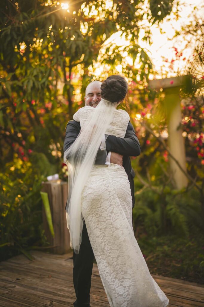 A groom embraces a bride in a white lace dress and veil outdoors, with sunlight filtering through trees in the background.