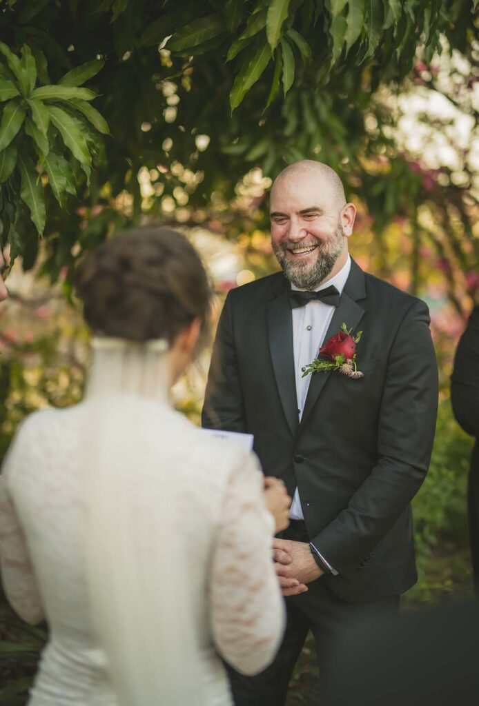 A groom in a black suit and bow tie smiles at a bride in a white dress and veil as she reads vows outdoors, with greenery in the background.