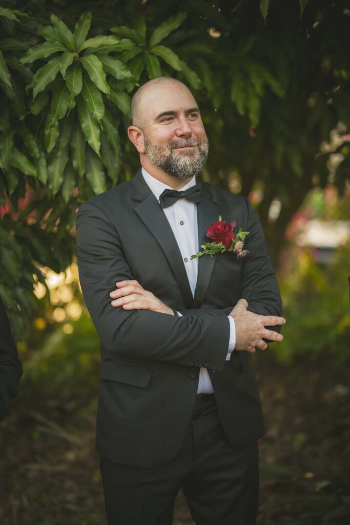 A man in a black tuxedo with a red boutonniere stands outdoors with arms crossed, smiling, in front of leafy green foliage.