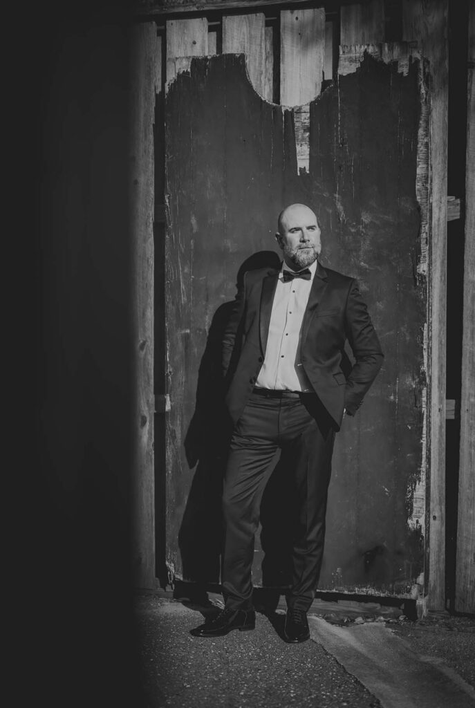 A man in a suit stands with his hands in his pockets against a weathered wooden door, looking to the side. The image is in black and white.