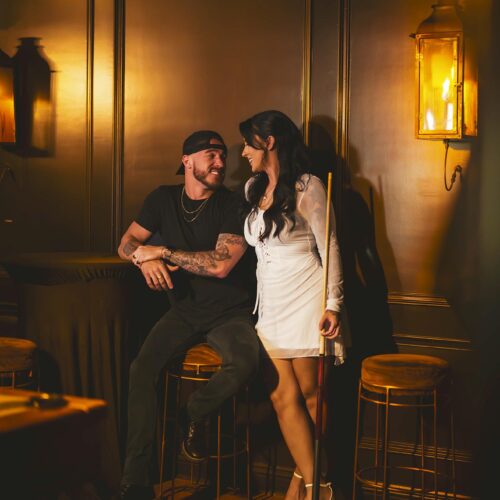A man sits on a barstool smiling at a woman standing next to him holding a pool cue in a warmly lit room with dark wood paneling.