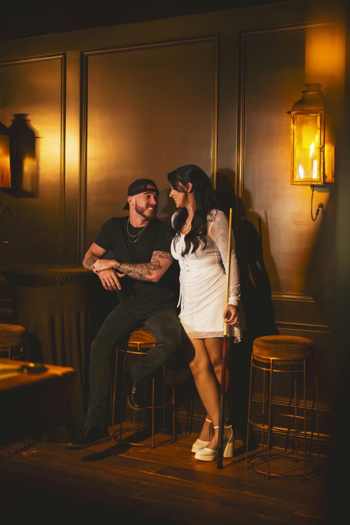 A man sits on a barstool smiling at a woman standing next to him holding a pool cue in a warmly lit room with dark wood paneling.