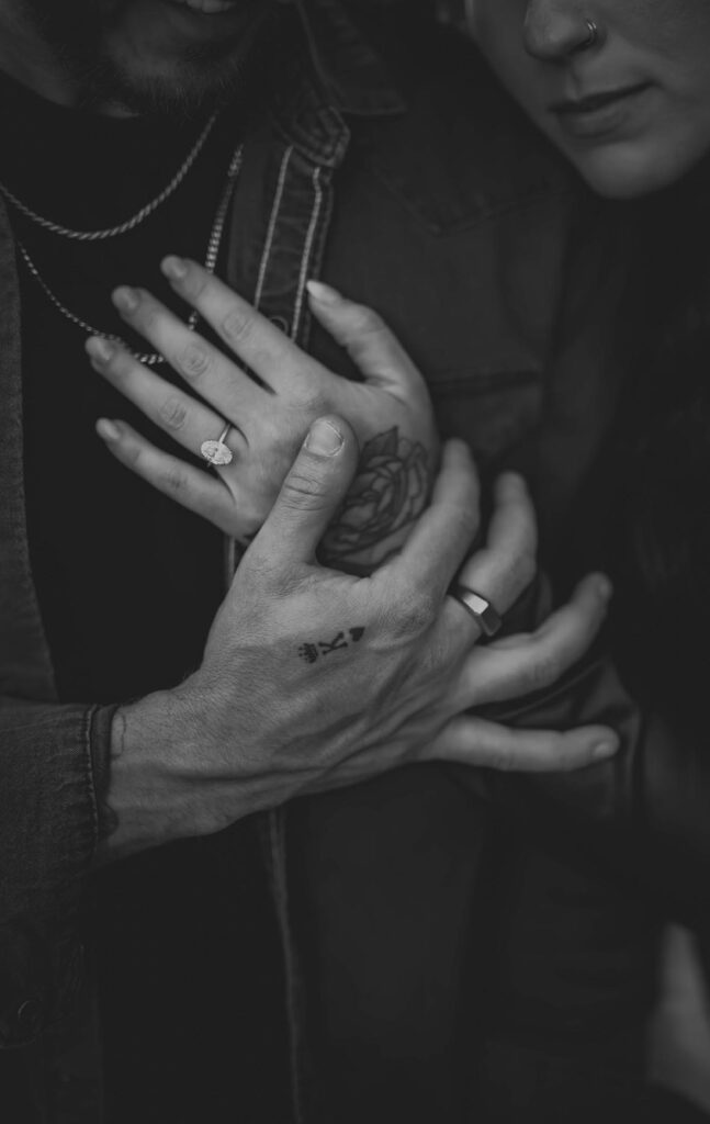 Close-up of two people’s hands intertwined, showing rings on their fingers and a tattoo on one hand, with both wearing dark clothing.
