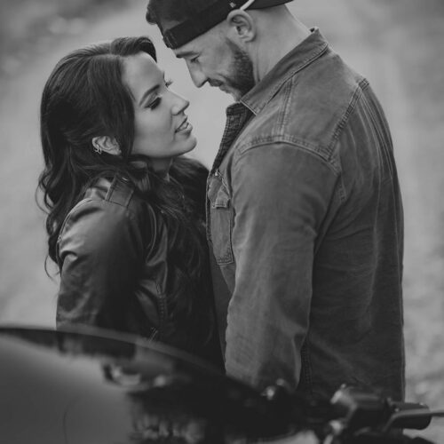 A man and woman stand closely facing each other on a dirt road, both wearing jackets. The man wears a backward cap. A motorcycle is visible in the foreground. The image is in black and white.