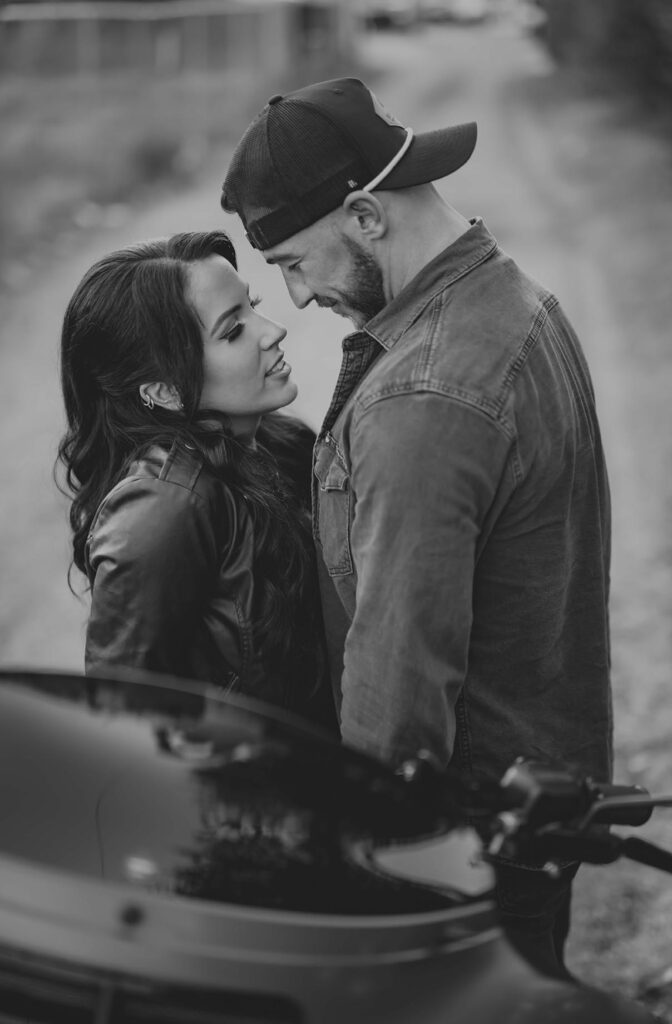 A man and woman stand closely facing each other on a dirt road, both wearing jackets. The man wears a backward cap. A motorcycle is visible in the foreground. The image is in black and white.