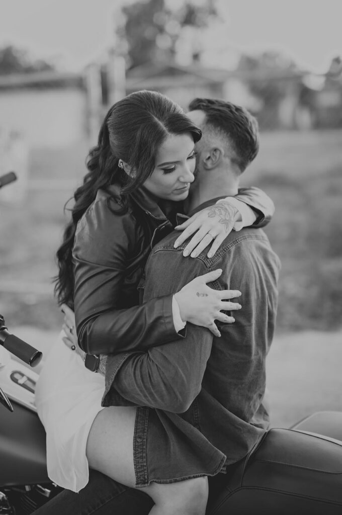 A woman with long hair hugs a man from behind while sitting together on a motorcycle outdoors. The image is in black and white.