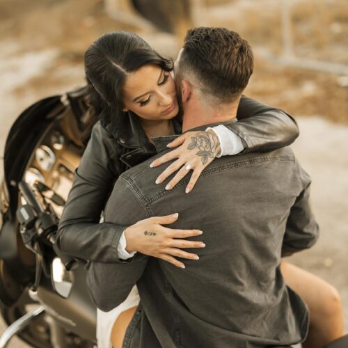 A woman sitting on a motorcycle embraces a man from behind, with her arms around his shoulders. They are outdoors on a dirt path with a fence and dry grass in the background.