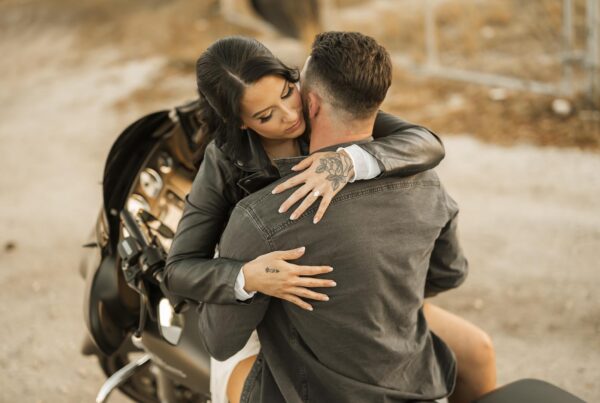 A woman sitting on a motorcycle embraces a man from behind, with her arms around his shoulders. They are outdoors on a dirt path with a fence and dry grass in the background.