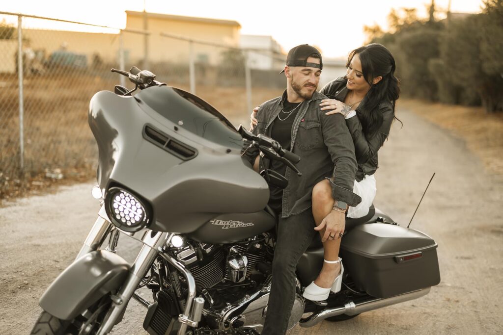 A man sits on a parked motorcycle while a woman, seated behind him, places her arms on his shoulders. Both are outdoors near a chain-link fence.