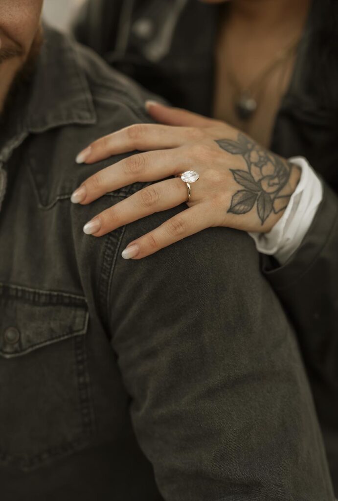 A person with a floral hand tattoo and an engagement ring rests their hand on another person’s shoulder, both wearing dark jackets.