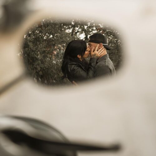 A couple is kissing, reflected in a motorcycle side mirror, with blurred foliage in the background.