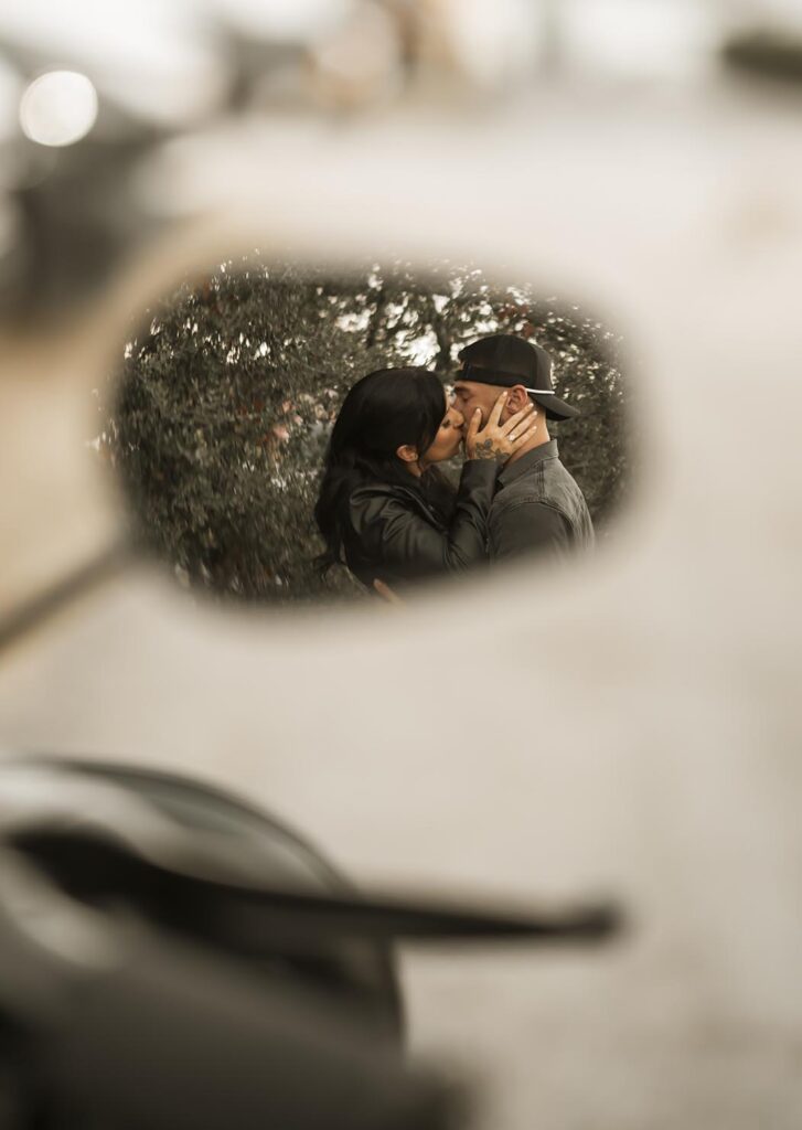 A couple is kissing, reflected in a motorcycle side mirror, with blurred foliage in the background.