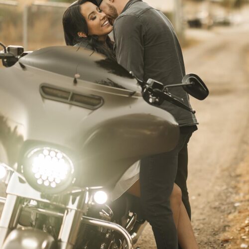 A man and woman smile and embrace beside a parked motorcycle on a sunlit outdoor path.