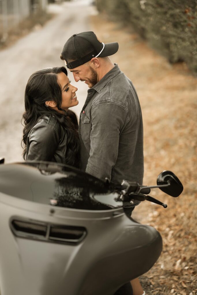 A man and woman stand close together smiling near a motorcycle on a path with dry leaves and bushes in the background.