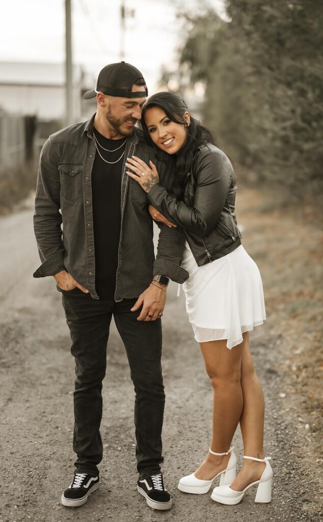 A man and woman stand together on a gravel path; she leans on his shoulder, both smiling. The man wears black clothes and a cap, the woman wears a black jacket and white dress with heels.