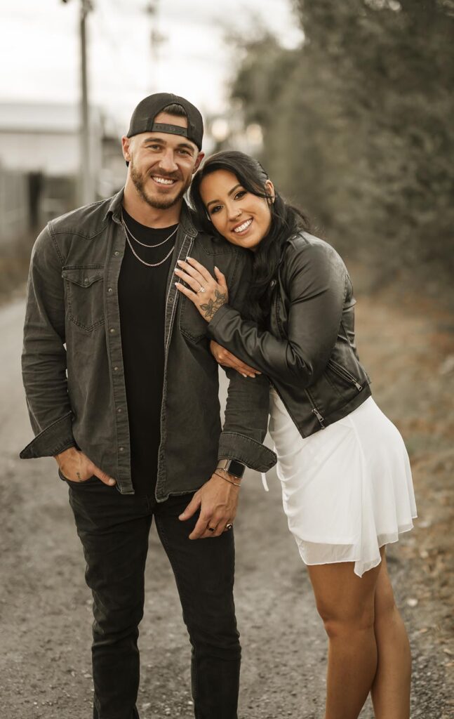 A man and woman stand close together on a gravel path; the woman leans on the man’s shoulder, both are smiling and dressed casually in jackets. Trees and buildings are visible in the background.