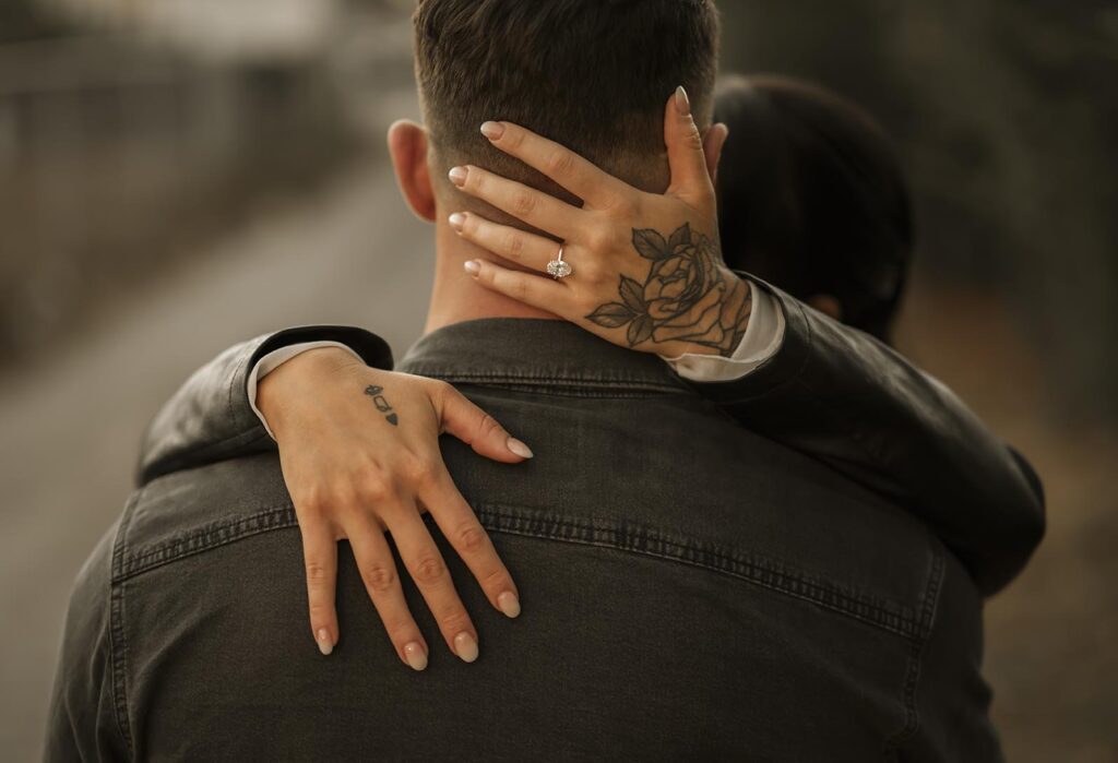 A person embraces another from behind, showing hands with a rose tattoo and an engagement ring, outdoors on a blurred path.