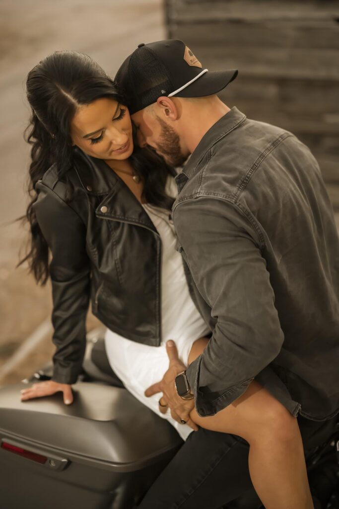 A man wearing a cap leans close to a woman seated on a motorcycle. Both wear jackets; he has his hand on her leg, and they appear to be sharing an intimate moment.